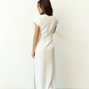 May include: A long, white dress with short sleeves and a high neckline. The dress has a side slit and is made of a smooth material. The model is seen from the back.