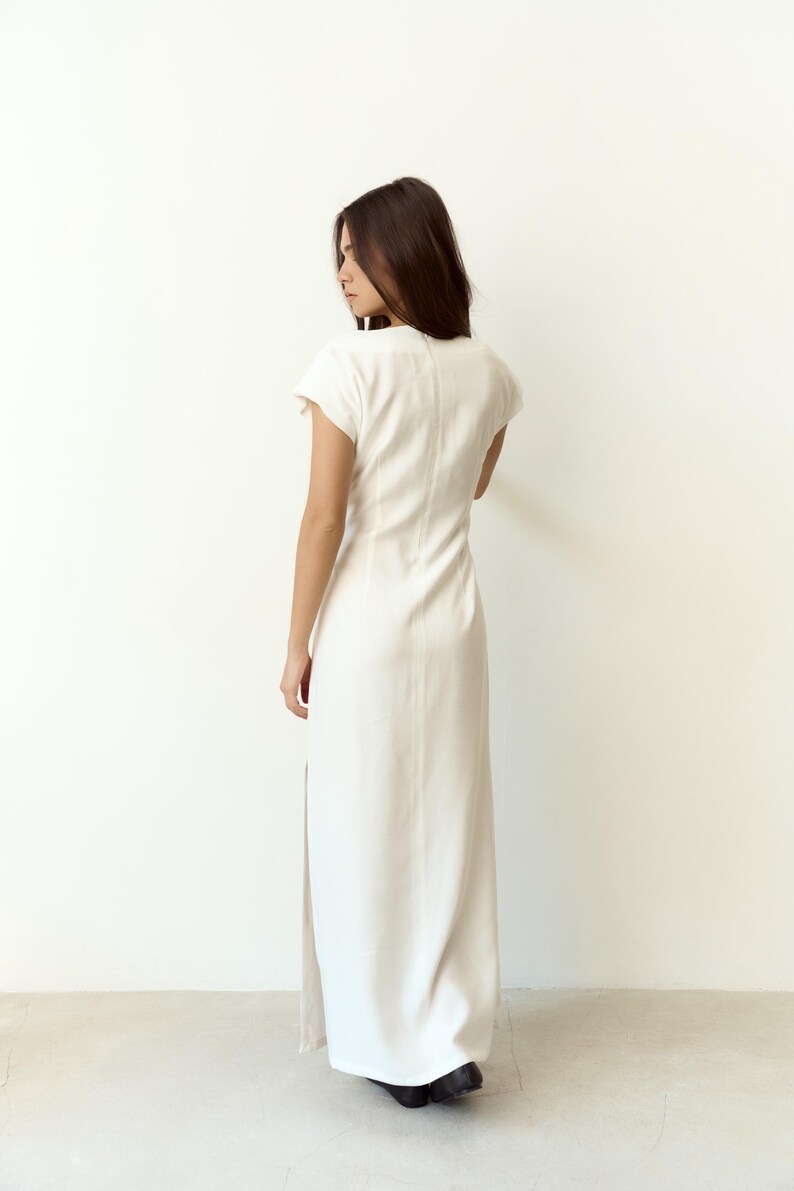 May include: A long, white dress with short sleeves and a high neckline. The dress has a side slit and is made of a smooth material. The model is seen from the back.