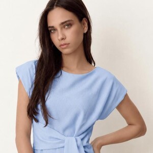 Puede incluir: Un vestido azul claro con cuello barco y mangas cortas. El vestido presenta un detalle de cintura anudada. La modelo de la imagen tiene el pelo largo y oscuro y lleva peque&ntilde;os pendientes de aro.