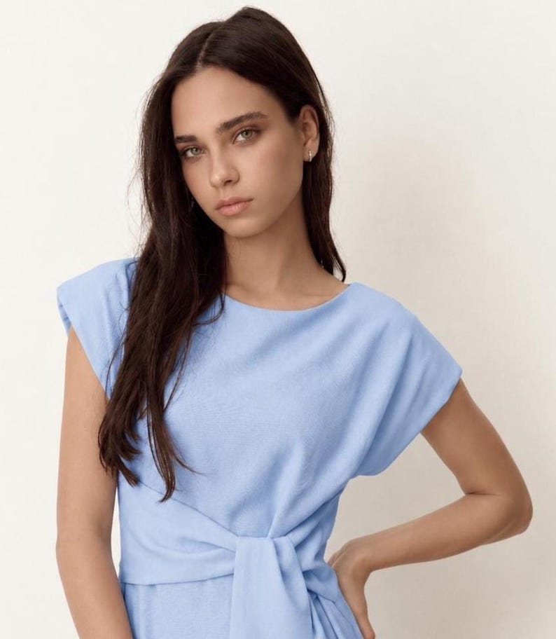 Puede incluir: Un vestido azul claro con cuello barco y mangas cortas. El vestido presenta un detalle de cintura anudada. La modelo de la imagen tiene el pelo largo y oscuro y lleva peque&ntilde;os pendientes de aro.