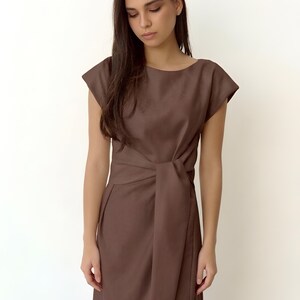 May include: A brown dress with a boat neckline and short sleeves. The dress has a wrap-style waist with a tie detail. The fabric appears to be a smooth, solid colour. The dress is knee-length.