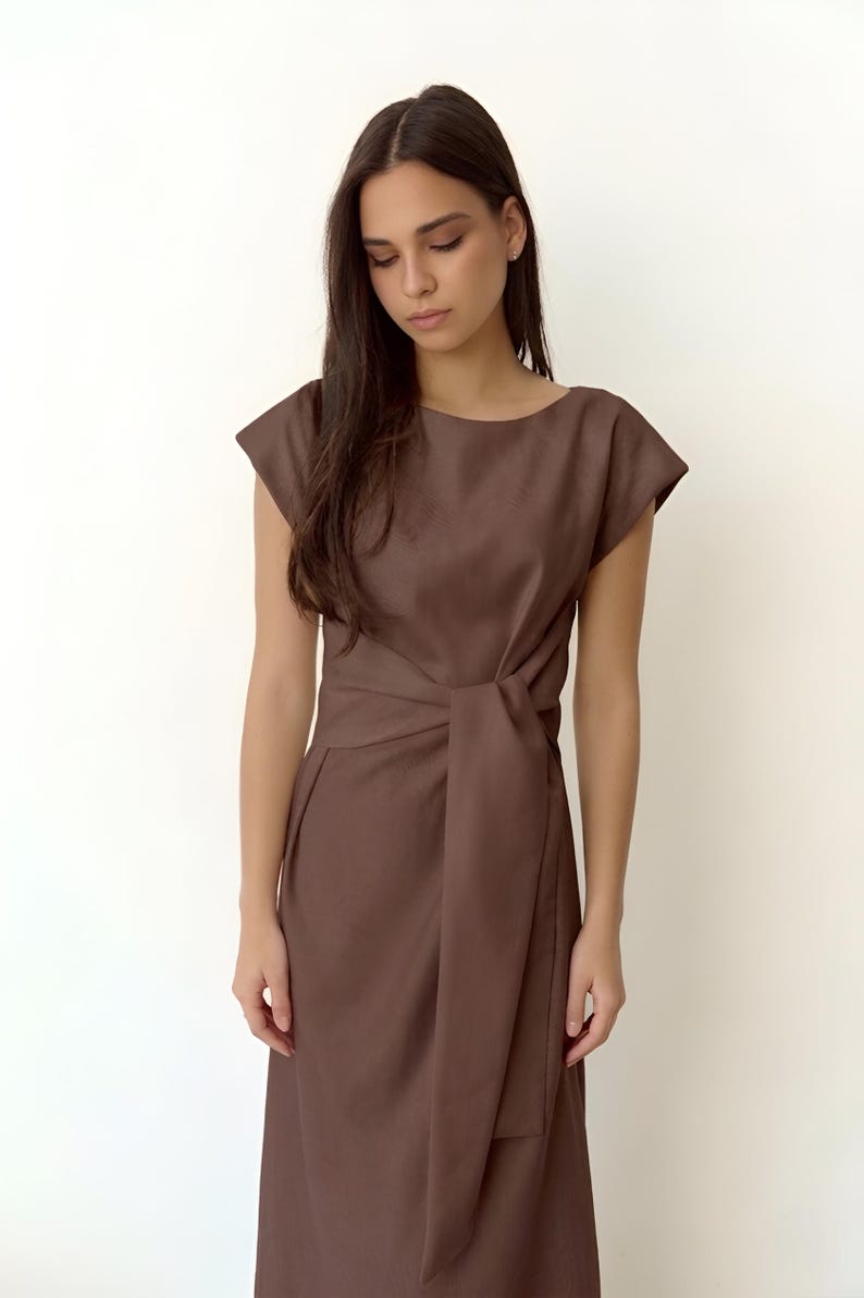 May include: A brown dress with a boat neckline and short sleeves. The dress has a wrap-style waist with a tie detail. The fabric appears to be a smooth, solid colour. The dress is knee-length.