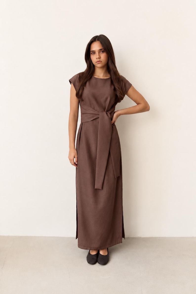 May include: A brown maxi dress with a tie-waist detail. The dress has short sleeves and a floor-length skirt with side slits. The model is wearing black flats. The dress is made of a solid, neutral-toned fabric.