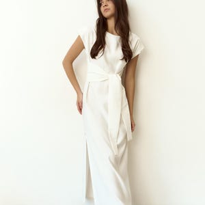 May include: A white dress featuring a tie-waist design and short sleeves. The dress has a side slit and is worn with black flats. The garment is made from a light fabric, suitable for multiple events. The dress is approximately 150 cm in length.