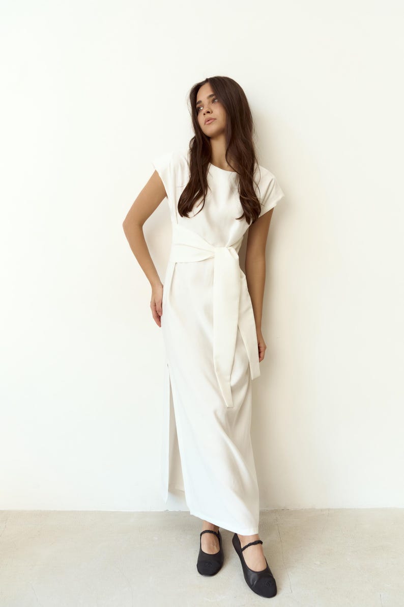 May include: A white dress featuring a tie-waist design and short sleeves. The dress has a side slit and is worn with black flats. The garment is made from a light fabric, suitable for multiple events. The dress is approximately 150 cm in length.