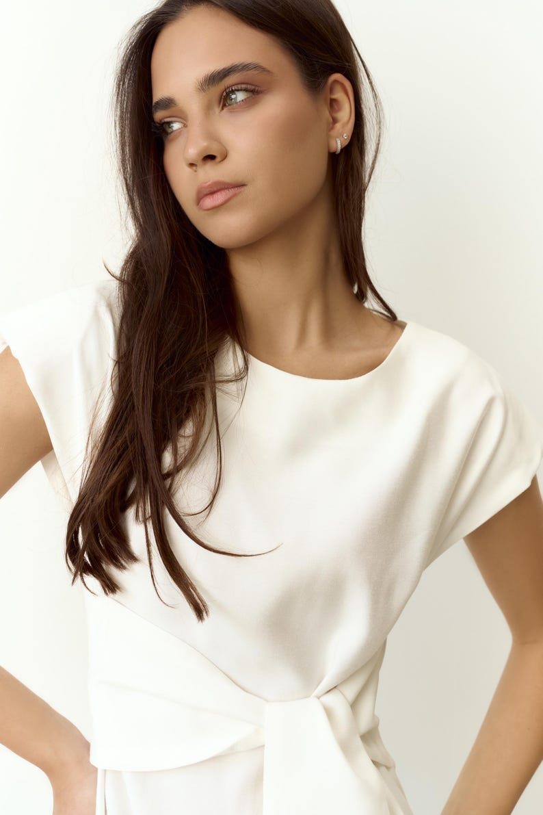 Puede incluir: Una blusa blanca de manga corta con cuello barco y cintura anudada. La blusa es de tejido liso. La persona que la lleva tiene el pelo oscuro y la piel clara. El estilo es minimalista y elegante.
