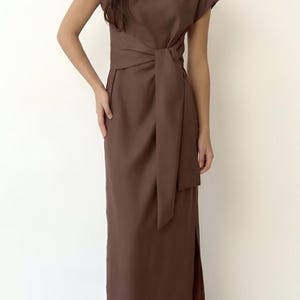 May include: A brown dress with cap sleeves, a knotted waist, and a floor-length skirt. The dress has a side slit and is made of a plain, matte fabric. The model is wearing black flats.