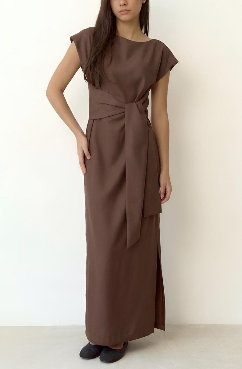 May include: A brown dress with cap sleeves, a knotted waist, and a floor-length skirt. The dress has a side slit and is made of a plain, matte fabric. The model is wearing black flats.