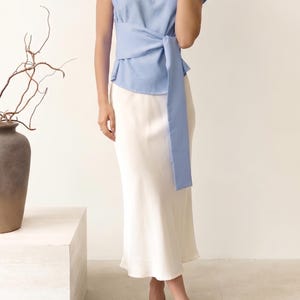Puede incluir: Una blusa azul claro con un lazo envolvente, combinada con una falda blanca. El conjunto se completa con tacones blancos de punta. La blusa tiene mangas cortas y una tela texturizada. La falda es de largo midi.