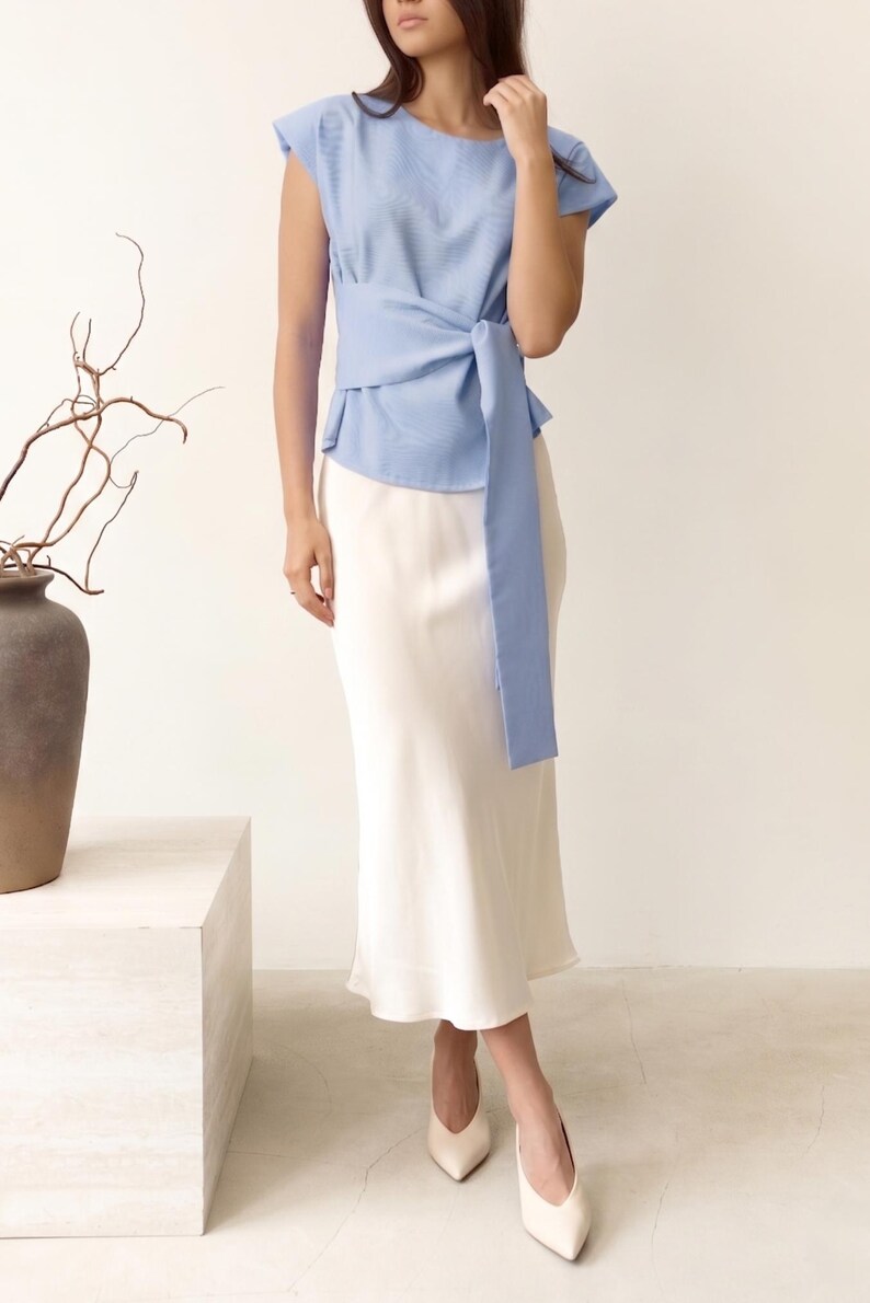Puede incluir: Una blusa azul claro con un lazo envolvente, combinada con una falda blanca. El conjunto se completa con tacones blancos de punta. La blusa tiene mangas cortas y una tela texturizada. La falda es de largo midi.