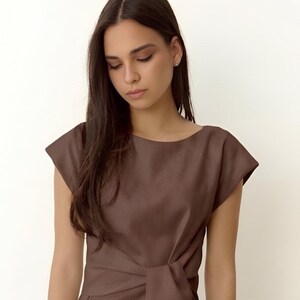 Puede incluir: Vestido marr&oacute;n con mangas cortas, cuello barco y detalle de nudo en la cintura. El vestido est&aacute; confeccionado con una tela lisa y de color liso. La modelo lleva el vestido.