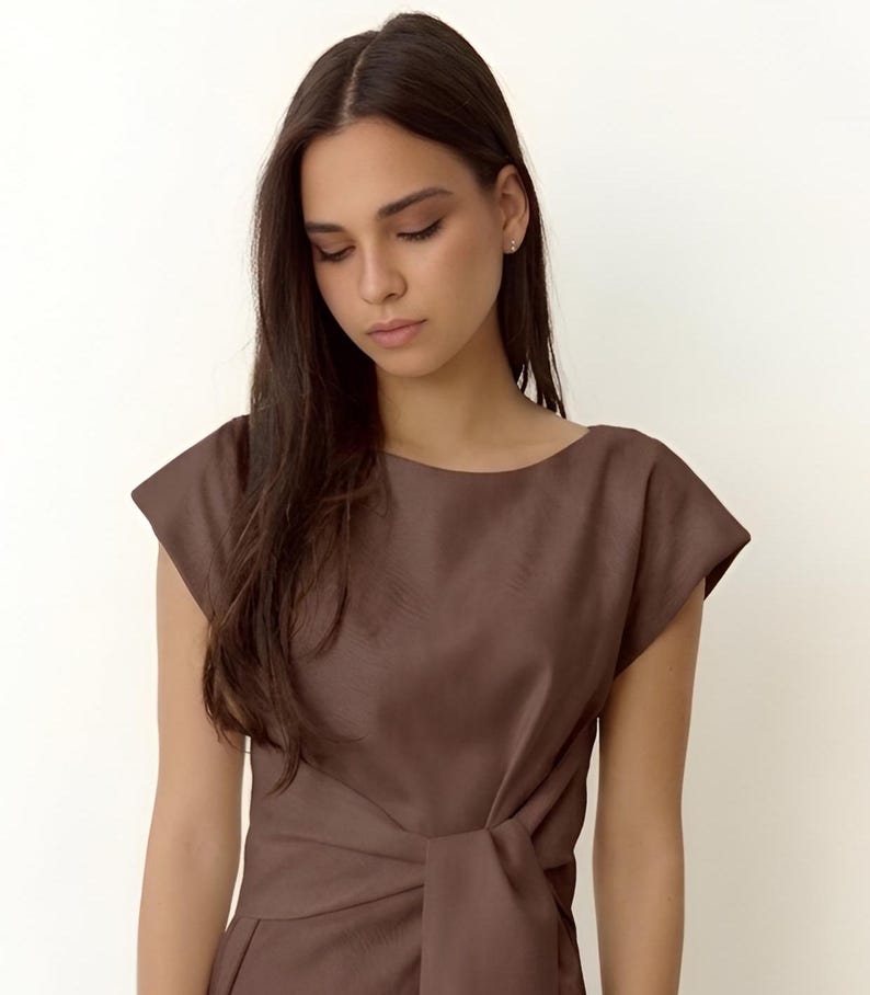 Puede incluir: Vestido marr&oacute;n con mangas cortas, cuello barco y detalle de nudo en la cintura. El vestido est&aacute; confeccionado con una tela lisa y de color liso. La modelo lleva el vestido.