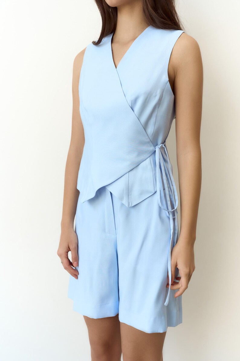 May include: A light blue sleeveless wrap top and matching shorts set. The top features a V-neck and tie closure. The shorts are high-waisted. The outfit is made of a smooth, solid-colored fabric.