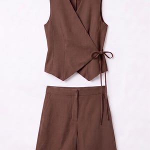 May include: A two-piece brown outfit featuring a sleeveless wrap top with a tie closure and matching shorts. The shorts have a tailored fit with front pockets. The outfit is made of a solid-colored fabric.