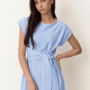 May include: A pale blue dress with a tie-waist. The dress has a round neckline and short sleeves. The model is wearing the dress. The dress is made of a soft, flowing fabric.