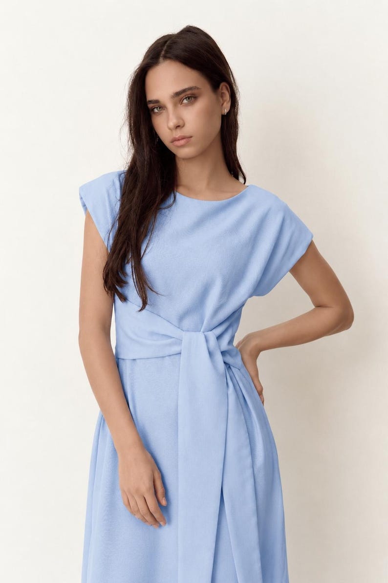 May include: A pale blue dress with a tie-waist. The dress has a round neckline and short sleeves. The model is wearing the dress. The dress is made of a soft, flowing fabric.
