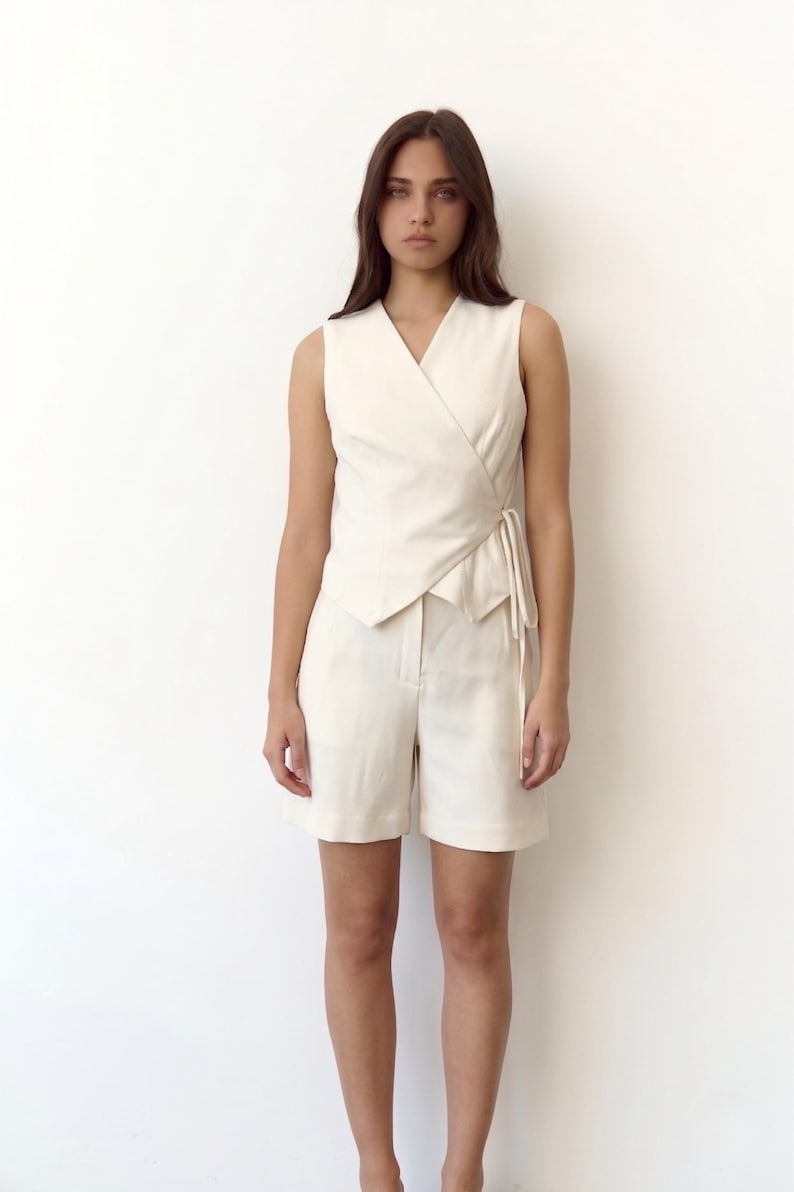 May include: A cream-colored sleeveless wrap top and matching shorts. The top has a V-neck and a tie closure at the waist. The shorts are high-waisted with a tailored fit. The outfit is made of a lightweight fabric.