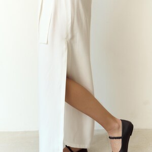May include: A person wearing a white, floor-length skirt with a high side slit. The person is wearing black ballet flats with an ankle strap. The flats have a small bow detail on the toe.
