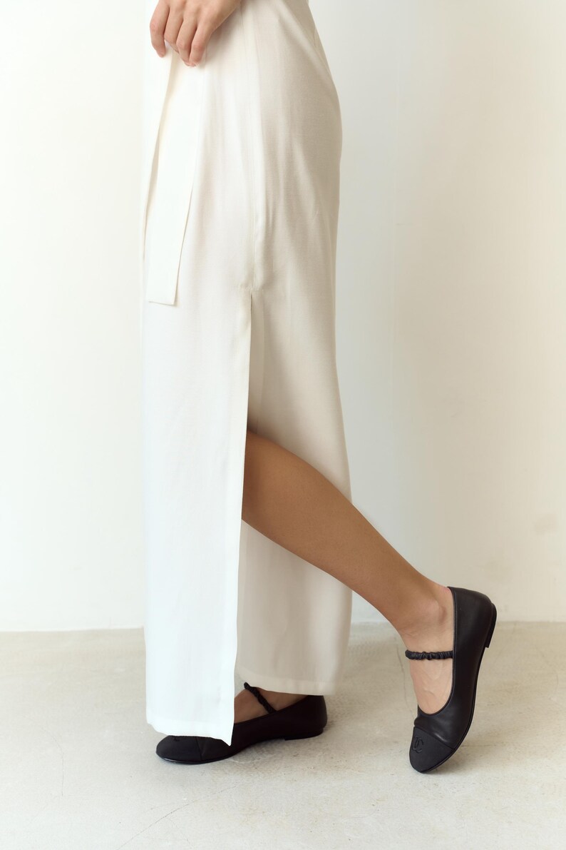 May include: A person wearing a white, floor-length skirt with a high side slit. The person is wearing black ballet flats with an ankle strap. The flats have a small bow detail on the toe.