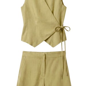 May include: A two-piece outfit in a light olive green color. The set includes a sleeveless wrap top with a tie closure and matching shorts. The fabric appears to be linen or a similar lightweight material, suitable for warm weather.