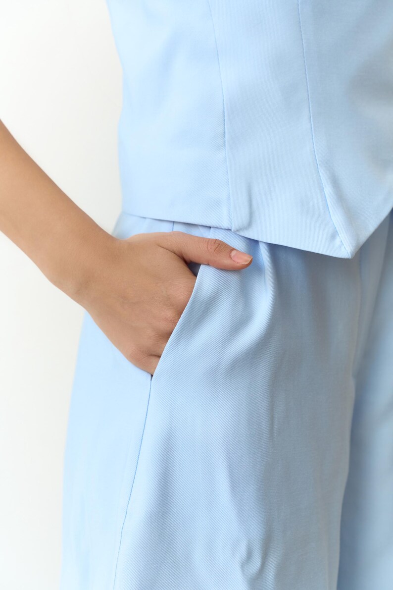 May include: Light blue two-piece outfit. The top has a tailored design, and the pants feature a pocket. The fabric appears to be a smooth, woven material. The outfit is a solid, pale blue color.