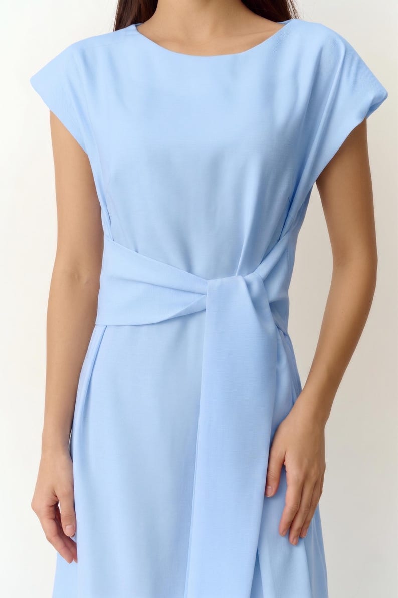 May include: A pale blue dress with short sleeves and a matching waist tie. The dress has a knotted detail at the waist. The fabric looks smooth and lightweight. The dress is a simple, elegant design.