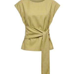 Puede incluir: Top de color verde oliva claro con una cinta envolvente en la cintura. La parte superior tiene mangas cortas y un cuello redondo. La corbata cuelga hacia abajo en el lado derecho. La tela parece ser un material suave similar al ante.