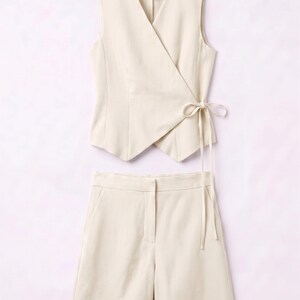 May include: A cream-colored two-piece outfit featuring a sleeveless wrap top with a tie closure and matching shorts. The ensemble is made of a smooth, solid-colored fabric, perfect for a coordinated look.