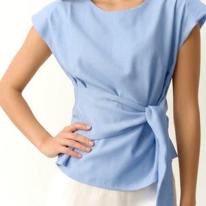 Puede incluir: Una blusa azul claro con un dise&ntilde;o envolvente. La blusa tiene mangas cortas y un detalle de nudo en la cintura. La tela parece ser un material suave y tejido. La modelo lleva una falda blanca.
