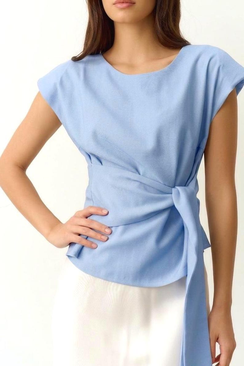 Puede incluir: Una blusa azul claro con un dise&ntilde;o envolvente. La blusa tiene mangas cortas y un detalle de nudo en la cintura. La tela parece ser un material suave y tejido. La modelo lleva una falda blanca.
