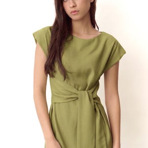 May include: Olive green dress with a boat neckline and cap sleeves. The dress has a wrap-style waist with a knotted detail. The model is wearing a ring. The dress is made of a soft, flowing fabric.