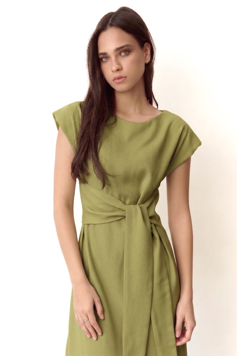 May include: Olive green dress with a boat neckline and cap sleeves. The dress has a wrap-style waist with a knotted detail. The model is wearing a ring. The dress is made of a soft, flowing fabric.