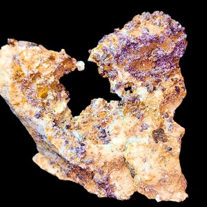 Pode incluir: Uma aproximação de um espécime mineral com um buraco no centro. A rocha tem uma superfície áspera e texturizada com tons de bege, castanho e roxo. O fundo é preto sólido.