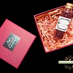 Op de afbeelding: Een rode geschenkdoos met bijpassende deksel, naast een open doos gevuld met versnipperd papier en een glazen fles. De fles bevat kleine, donkerrode kristallen en een label met tekst. De dozen staan op een zwarte achtergrond.