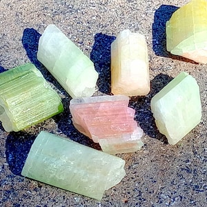 Peut inclure: Une collection de sept cristaux naturels polis, verts, jaunes et roses. Les cristaux sont de formes et de tailles différentes et sont disposés de manière aléatoire.