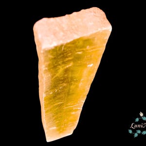 Pode incluir: Um cristal de calcita bruto, dourado, com uma superfície áspera e texturizada. O cristal tem uma forma retangular com uma borda ligeiramente angulada. O fundo é preto, destacando a cor e a textura do cristal. O logotipo LuniNature está no canto inferior direito.