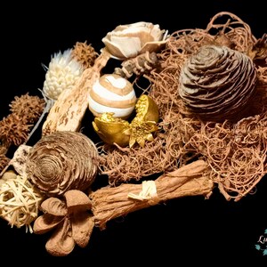 Puede incluir: Un surtido de elementos decorativos naturales sobre un fondo negro. El arreglo incluye flores secas, piñas, vainas de semillas y esferas tejidas en tonos marrón, beige y dorado. Un elemento decorativo dorado está en el centro.