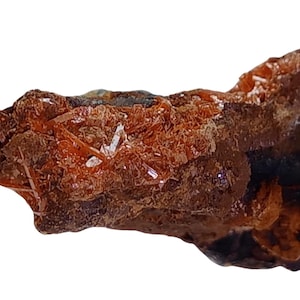 Könnte beinhalten: Nahaufnahme eines rauen, braunen Gesteins mit eingebetteten orangefarbenen Kristallen. Die Kristalle sind zusammen gruppiert und haben ein glänzendes, metallisches Aussehen.