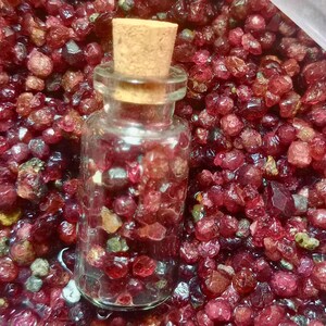 May include: A glass bottle filled with small, red, ruby-colored gemstones. The bottle has a cork stopper and is sitting on a bed of loose, red gemstones.