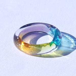 May include: A clear resin ring with a gradient of colors, including purple, yellow, green, and blue. The ring is round and has a smooth, polished surface. The ring is casting a shadow on a white surface.