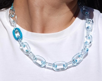 Puede incluir: Un collar y pendientes a juego de cadena acrílica transparente con detalles en azul claro. El collar presenta eslabones ovalados grandes, con un solo eslabón azul. Los pendientes tienen un diseño similar, con una pequeña cuenta azul.