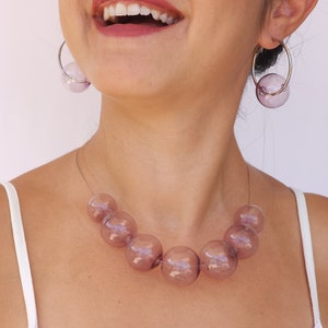 May include: A mauve glass bead necklace with large, round beads. The necklace is worn by a person with a white tank top and hoop earrings with mauve glass beads.