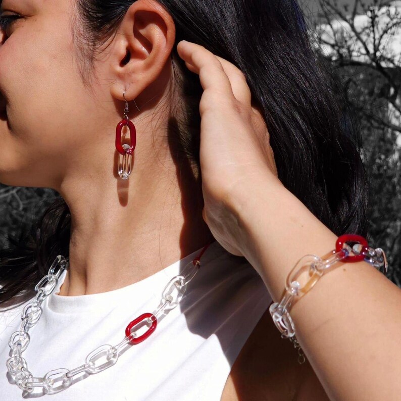 May include: Clear chain-link necklace, bracelet, and earrings with red oval accents. The jewelry is made of transparent material, creating a modern and minimalist aesthetic. The earrings hang from silver hooks.