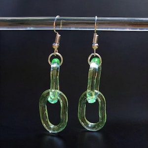 May include: A pair of handmade earrings featuring a chain link design. The earrings are made of translucent, light green glass. The chain links are connected with silver-colored metal findings and a small green bead. The earrings hang from silver-colored ear wires.