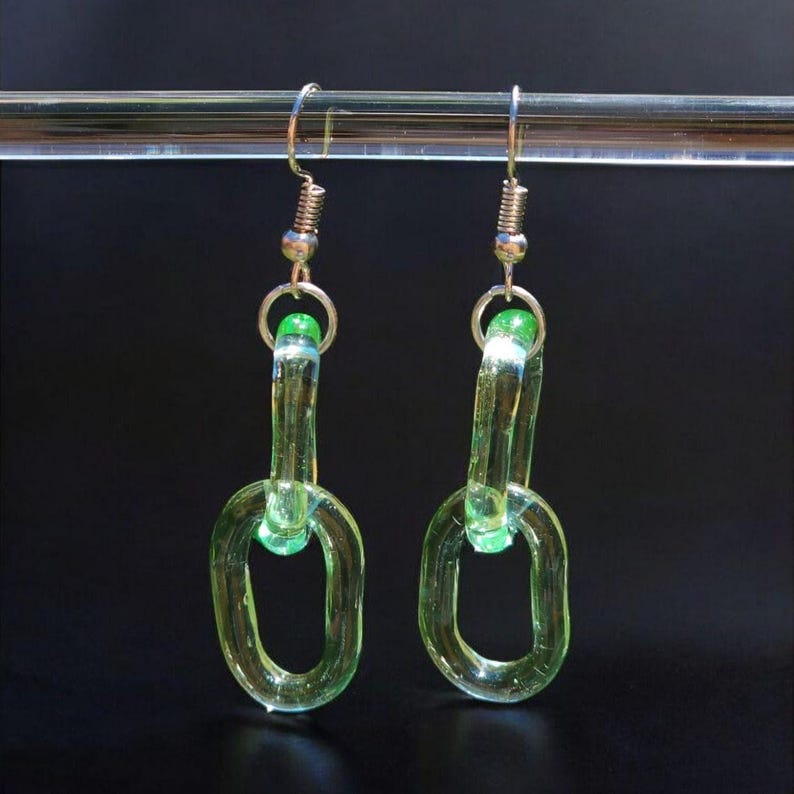 May include: A pair of handmade earrings featuring a chain link design. The earrings are made of translucent, light green glass. The chain links are connected with silver-colored metal findings and a small green bead. The earrings hang from silver-colored ear wires.