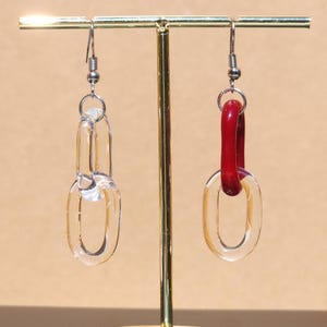 May include: A pair of clear acrylic chain link earrings. One earring is entirely clear, while the other features a red acrylic link. The earrings hang from silver-colored ear wires. The earrings are displayed on a gold-colored stand.