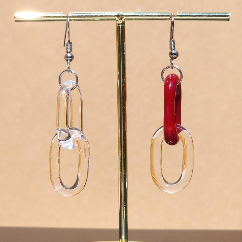 May include: A pair of clear acrylic chain link earrings. One earring is entirely clear, while the other features a red acrylic link. The earrings hang from silver-colored ear wires. The earrings are displayed on a gold-colored stand.
