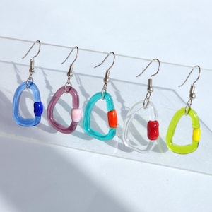 Peut inclure: Cinq boucles d'oreilles en résine transparente colorées. Chaque boucle d'oreille est d'une couleur différente : bleu, violet, turquoise, transparent et vert. Chacune a une petite couleur d'accentuation contrastante, comme le rouge ou le jaune, et des crochets d'oreilles argentés.