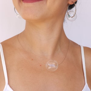May include: A woman wearing a white vest top with a silver necklace featuring a large clear glass bubble pendant. She is also wearing silver hoop earrings with clear glass bubble charms.