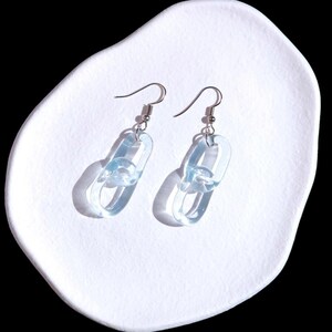May include: A pair of light blue chain link earrings with silver-colored hooks. The earrings are made of a translucent material and are displayed on a white, organically shaped surface.